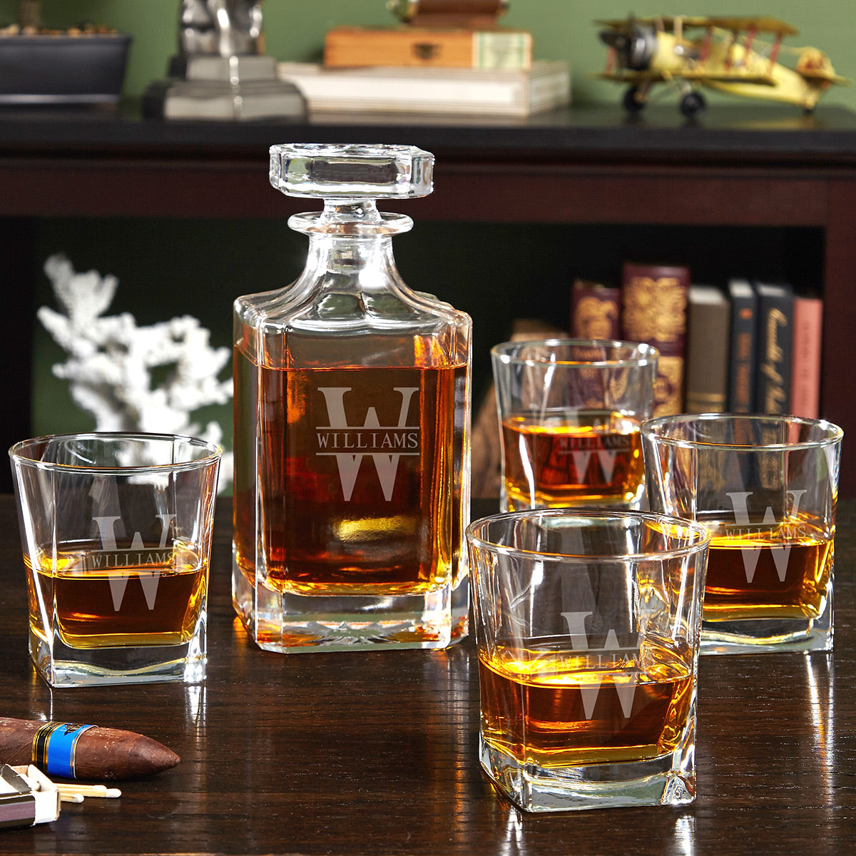 Glass decanter and four whiskey tumblers with “WILLIAMS W” monogram on a wooden table, books behind.