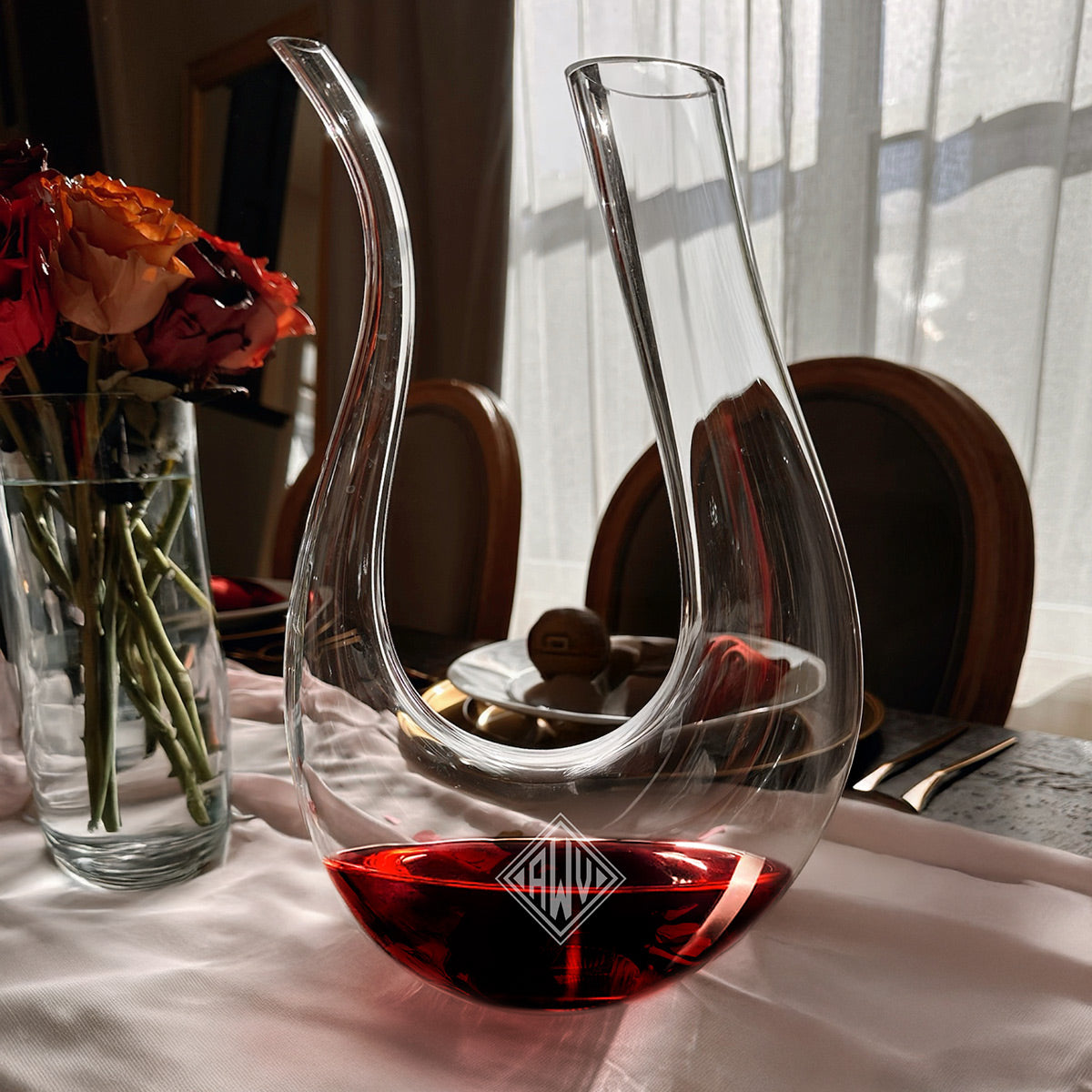 Clear glass decanter with red wine on a table beside a vase of roses, with chairs and window curtains behind.
