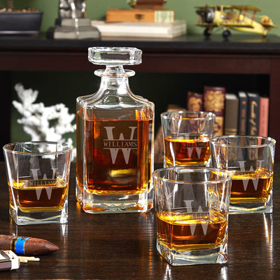 Glass decanter and four whiskey tumblers with “WILLIAMS W” monogram on a wooden table, books behind.