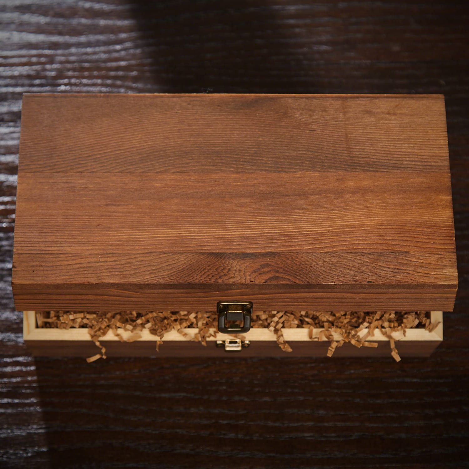 Wooden box with metal latch, slightly open, showing crinkle paper filler inside on a dark surface.